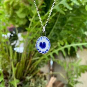 Puede incluir: Un collar de plata con un colgante circular. El colgante presenta una base blanca con un patrón de teñido anudado azul. El collar se muestra sobre un fondo de follaje verde.