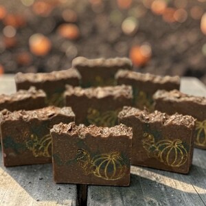 May include: A collection of rectangular brown soap bars arranged in a cluster. Each bar features a textured top and a gold-colored graphic print of a pumpkin with vine details on the front face. The soap bars have a dark, earthy brown color.