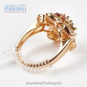 Puede incluir: Un anillo de oro con una montura de piedras preciosas multicolores. El anillo está en un ajustador de tamaño de anillo transparente.