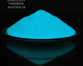 May include: A pile of bright blue glow-in-the-dark powder on a black surface. The powder is in a small glass dish.