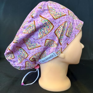 May include: A lavender scrub cap featuring a repeating pattern of triangular pastries topped with colourful sprinkles. The cap includes a white elastic cord with a small pink toggle for adjustment and a white fabric tag labelled ScrubbiesCo.