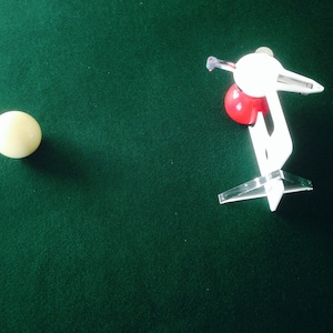 May include: A white billiard ball on a green felt pool table with a white cue stick and a red ball on a white ball return device.