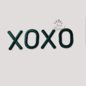 Könnte beinhalten: Dunkelgrüne gestickte Buchstaben, die XOXO buchstabieren, mit einer texturierten, erhabenen Oberfläche. Über den Buchstaben befindet sich ein kleines graues Logo mit einem Sonnenmotiv und dem Text Sun Kissed. Die Buchstaben haben abgerundete Kanten.