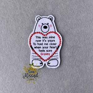 Może przedstawiać: Biała filcowa naszywka w kształcie misia z czerwonym sercem. Serce zawiera tekst "This was mine now it's yours To hold me close when your heart feels sore Granny". Miś ma czarny nos i oczy oraz różowe policzki.