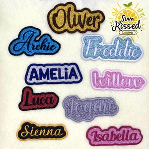 Könnte beinhalten: Eine Sammlung von zehn personalisierten Namensaufnähern aus Glitzerstoff in verschiedenen Farben wie Gold, Blau, Rot, Pink und Lila. Jeder Aufnäher zeigt einen einzigartigen gestickten Namen wie Oliver, Archie, Freddie, Amelia, Willow, Luca, Logan, Sienna und Isabella.