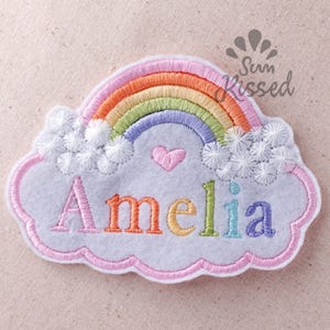 Puede incluir: Un parche bordado en forma de nube gris claro con un arcoíris colorido, nubes blancas bordadas, un pequeño corazón rosa y el nombre Amelia bordado en letras multicolores. El parche tiene un borde cosido en color rosa.