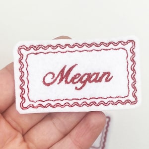 May include: White felt name tag embroidered with the name "Megan" in red script. The tag features a red, scalloped border. The rectangular name tag is held in a person's hand.