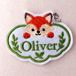 Pode incluir: Um emblema de feltro branco de formato oval com uma cabeça de raposa bordada em laranja e creme no topo. O nome Oliver está bordado em letras verdes abaixo da raposa, ladeado por ramos de folhas verdes. O texto Sun Kissed aparece no canto superior direito.