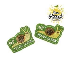 May include: Two embroidered patches featuring a snail design with the words "SLOW DOWN". The snails have yellow bodies and brown shells, set against a green background. A "Sun Kissed Lemon" logo is in the upper right corner.