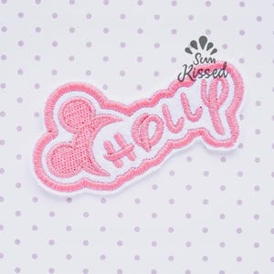Könnte beinhalten: Ein gestickter Aufnäher in Pink und Weiß mit dem Namen Holly in Schreibschrift. Das Design enthält links eine stilisierte Silhouette von Mausohren, wobei die gesamte Form von einem dicken pinkfarbenen Rand auf weißem Grund umgeben ist.