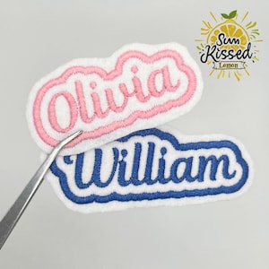 Peut inclure: Deux écussons brodés avec des noms. L'un présente le nom "Olivia" en fil rose avec un contour rose, et l'autre le nom "William" en fil bleu avec un contour bleu. Les deux sont sur un fond blanc.