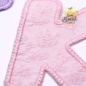 May include: A light pink fabric letter K patch featuring embroidered floral patterns and a stitched border. The patch displays the text Sun Kissed Lemon in a circular logo design with a yellow lemon graphic and green leaves at the top.