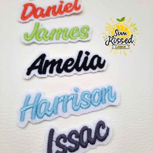 Op de afbeelding: Geborduurde naamlabels in verschillende kleuren: rood "Daniel", groen "James", zwart "Amelia", lichtblauw "Harrison" en donkerblauw "Issac". Elk label heeft een witte rand. Een "Sun Kissed Lemon" logo is ook zichtbaar.