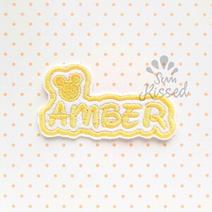 Può includere: Una toppa ricamata che presenta il nome AMBER in filo giallo su sfondo bianco con un bordo giallo. Sopra il nome c'è una piccola sagoma di testa di topo ricamata in giallo. La toppa ha un bordo smerlato e cuciture gialle.