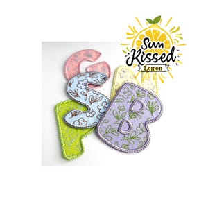 May include: Embroidered fabric letters in various colors, including pink, blue, green, and purple, with floral and leaf patterns. The letters spell out "SBMP". The image also includes a logo that says "Sun Kissed Lemon".