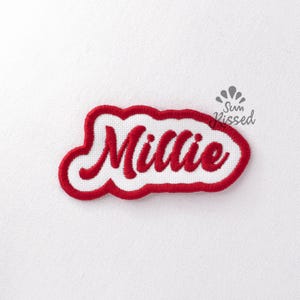 May include: A white fabric patch featuring the name Millie embroidered in red cursive script. The patch has a thick red embroidered border that follows the irregular shape of the lettering. Above the name, the text Sun Kissed is printed.