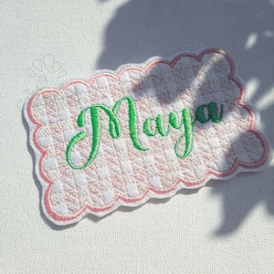Könnte beinhalten: Ein rechteckiger Stickaufnäher mit gewelltem Rand und einem rosa-weiß karierten Muster. Der Name Maya ist mittig in einer leuchtend grünen Schreibschrift aufgestickt. Der Aufnäher ist mit einem dicken rosa Rand versehen.
