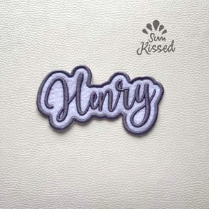 May include: A custom embroidered patch featuring the name Henry in a dark purple cursive script font. The lettering is outlined in a matching dark purple border and set against a white felt background. The text Sun Kissed is visible above.