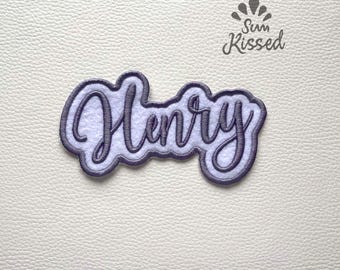 May include: A custom embroidered patch featuring the name Henry in a dark purple cursive script font. The lettering is outlined in a matching dark purple border and set against a white felt background. The text Sun Kissed is visible above.