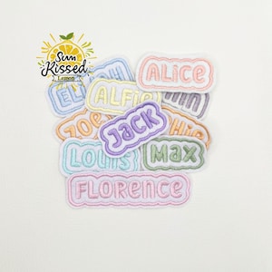 Puede incluir: Colección de parches bordados con nombres en varios colores pastel, incluyendo rosa, azul y verde. Cada parche presenta un nombre diferente, como "Alice" y "Jack", en un color contrastante. Los parches están dispuestos sobre una superficie blanca.