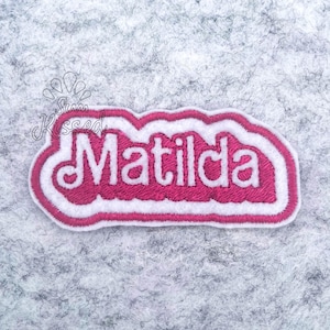 May include: A fabric patch featuring the name Matilda embroidered in white thread with a thick pink border. The text is centred on a pink background with a white outline. Above the name, the text Sun Kissed is printed in a thin black font.