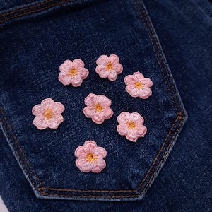 Könnte beinhalten: Sieben kleine, fünfblättrige rosa Blumen mit gelben Mitten sind auf eine dunkelblaue Jeanstasche gestickt. Die Stickerei besteht aus hellrosa Garn für die Blütenblätter und goldgelbem Garn für die Mitte jeder einzelnen Blume.