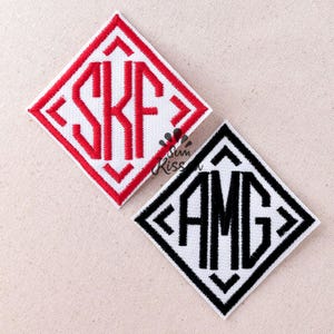 Op de afbeelding: Twee ruitvormige geborduurde patches op een lichte stof. De ene patch heeft een rode rand en het monogram SKF in rood garen. De tweede patch heeft een zwarte rand en het monogram AMG in zwart garen. Beide hebben een witte achtergrond.