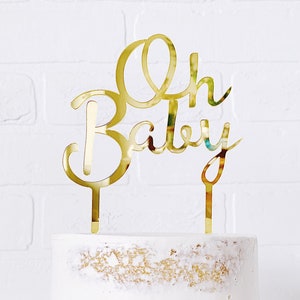 May include: Gold cake topper with the words "Oh Baby" in a cursive font. The topper is on top of a white cake with a light brown layer.