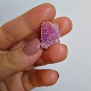 May include: A rough-cut, raw pink gemstone with a crystalline structure. The gemstone is held between fingers, showcasing its natural, irregular shape and vibrant color. The stone has a slightly translucent quality.