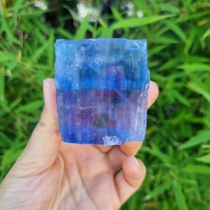 May include: A rectangular block of translucent blue and purple mineral with a crystalline, textured surface. The object displays distinct horizontal color bands, transitioning from a deep blue at the top to a violet-purple hue in the center section.