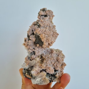 May include: A mineral specimen featuring a cluster of pale pink, textured crystalline formations interspersed with dark metallic gray mineral deposits and translucent, sharp, clear quartz points at the base of the structure.