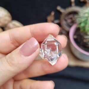 Pode incluir: Um cristal de quartzo transparente de terminação dupla com formato hexagonal e extremidades pontiagudas. A pedra apresenta inclusões internas e facetas naturais que refletem a luz, exibindo uma aparência transparente e vítrea com bordas geométricas.