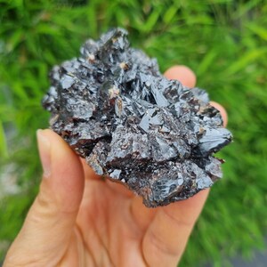 Peut inclure: Un spécimen minéral brut, sombre et brillant, composé d'un amas de cristaux pointus, anguleux et à multiples facettes. La surface de la pierre présente une coloration noire profonde avec des reflets métalliques et des inclusions brun-rouge.