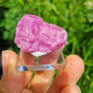 May include: A vibrant pink, crystalline mineral specimen with a rough, multifaceted texture. The stone is mounted on a small, square, clear acrylic base, showcasing its natural, jagged edges and deep magenta coloration in a raw, unpolished state.