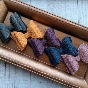 May include: Six handmade fabric hair bows in various colors, including black, mustard yellow, purple, teal, and pink. The bows are arranged on a gold tray with a beaded border.