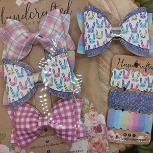 May include: Set of four handmade hair bows featuring a pink and white plaid bow, a pink and white gingham bow, and two bows with a white background and a pattern of colorful bunnies. The bows are displayed on a brown background with the text "Handcrafted" and "@handcraftedhenryave".