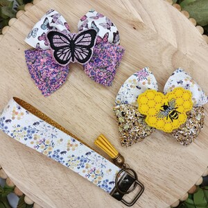 May include: Two fabric hair bows with a floral and butterfly design and a bee and honeycomb design. A white and yellow floral patterned key chain with a gold tassel.