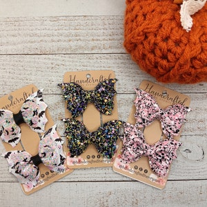 May include: Three sets of handmade hair bows with bat wings. The bows are made with black and white glitter fabric with a black center. The wings are white with a black bat silhouette. The bows are packaged on a card with the text "Handcrafted by Avenue".