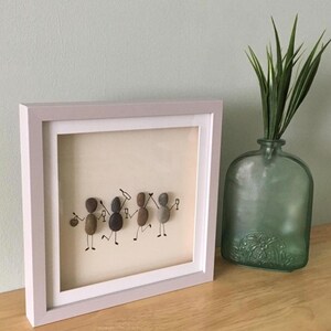 May include: A framed artwork featuring pebble art figures in a party scene. The figures are holding wine glasses and bottles, with black marker details. The frame is white, and the background is a light beige color. A green glass vase with plants is next to the frame.