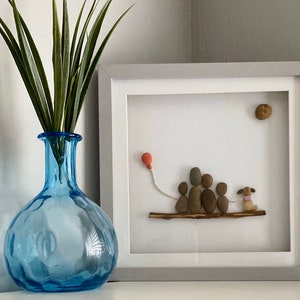 Op de afbeelding: Een ingelijst kunstwerk van kiezelsteen dat een gezin van vijf met een hond laat zien die op een tak loopt. Het gezin houdt een rode ballon vast. De achtergrond is wit met een bruine kiezelsteen in de rechterbovenhoek.