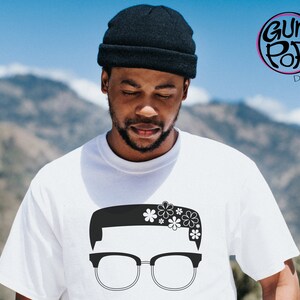 May include: A white t-shirt with a black and white graphic of a person with glasses and flowers in their hair. The graphic is a silhouette of a person with a short haircut and glasses. The flowers are stylized and have five petals each.
