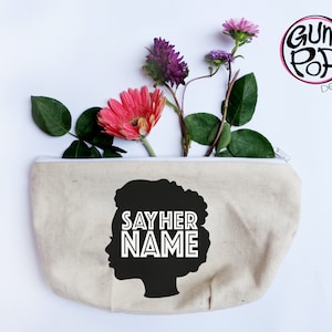 May include: A beige canvas zippered pouch with a black silhouette of a woman's profile and the words "SAY HER NAME" in white. The pouch is filled with pink, purple, and green flowers. The Gum Pop Designs logo is in the upper right corner.