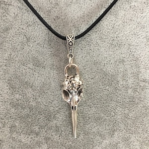 May include: Silver-toned pendant necklace featuring a bird skull design. The pendant hangs from a black cord and has intricate detailing on the skull and the bail. The background is a solid gray.