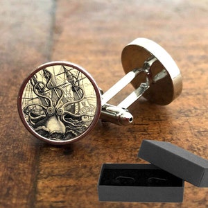 Può includere: Gemelli d'argento con un'immagine in bianco e nero di un polpo che attacca una nave. I gemelli sono presentati in una scatola regalo nera.