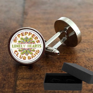 May include: Silver cufflinks with a circular face displaying the text "Sgt. Peppers Lonely Hearts Club Band" in a vibrant design. The cufflinks are presented in a black gift box, ideal for a music enthusiast.