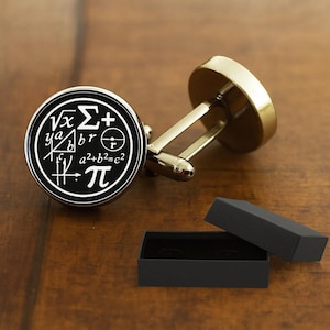 May include: A pair of gold-toned cufflinks with a black face featuring white mathematical symbols and equations. The cufflinks are round and have a classic design. A black box is open next to the cufflinks.