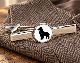 Könnte beinhalten: Eine silberfarbene Krawattenklammer mit einem runden Akzent, der die schwarze Silhouette eines Spaniels auf weißem Hintergrund zeigt. Der Metallclip hat eine polierte Oberfläche und einen Federmechanismus zur Befestigung an der Krawatte.