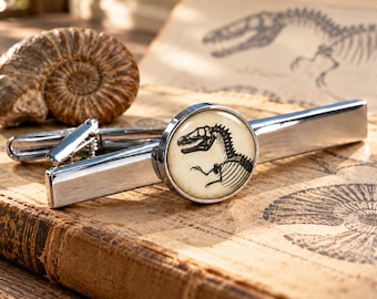 Könnte beinhalten: Eine silberfarbene Metall-Krawattenklammer mit einer runden Glaskabochon-Einlage. Die Einlage zeigt eine schwarz-weiße Illustration eines Dinosaurierskeletts auf cremefarbenem Grund. Die Krawattenklammer hat eine polierte, reflektierende Oberfläche.
