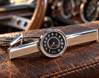 Könnte beinhalten: Eine silberfarbene Krawattenklammer mit einem runden Tachometer-Zifferblatt. Das schwarze Zifferblatt zeigt weiße Zahlen von 0 bis 120, das Wort MILES und einen dünnen orangefarbenen Zeiger. Der Metallclip hat eine polierte Oberfläche und Feder.