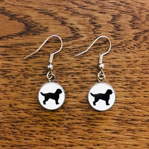 May include: A pair of silver dangle earrings with a black silhouette of a dog on a white round charm.